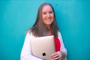 A smiling woman holding a laptop in front of a blue background.